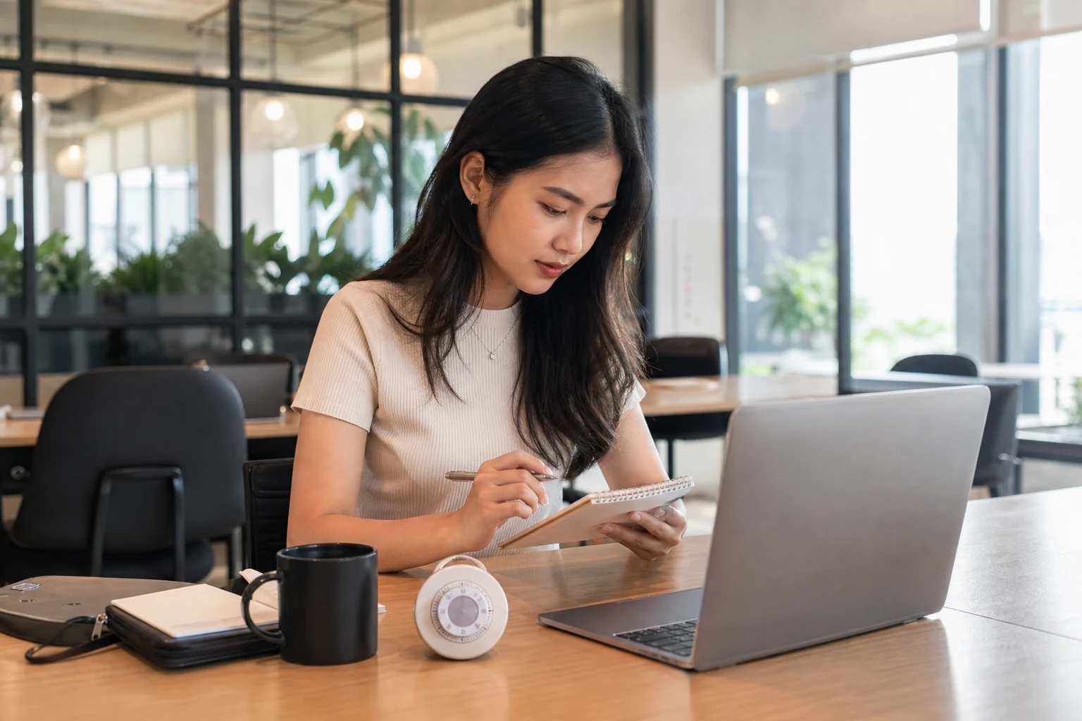 Seorang perempuan muda Chinese Indonesian meninjau daftar tugas di samping laptopnya di meja kerja bersama yang terang.